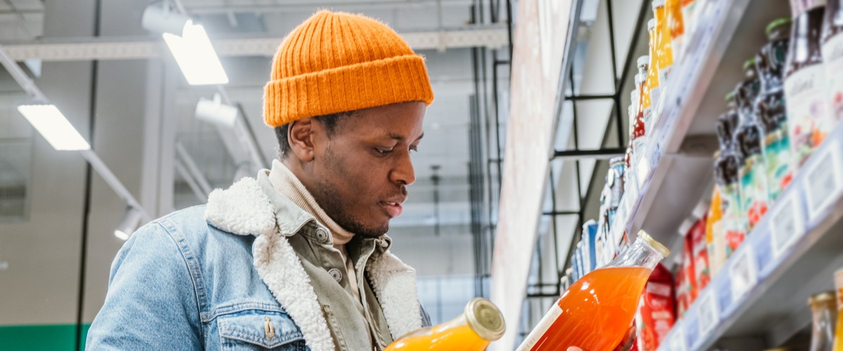 African man in a stylish orange hat and denim jacket chooses natural juice in glass bottles in a grocery supermarket standing at the shelves with products